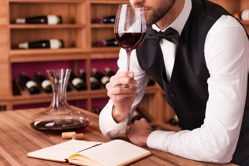 Sommelier examining wine. Cropped image of confident male sommelier examining wine while smelling it and leaning at the wooden table with wine shelf in the background